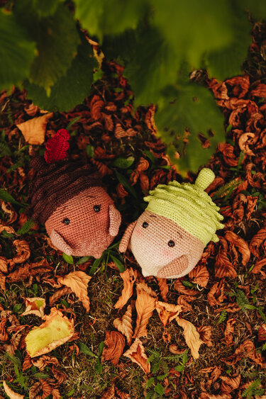 Zwei gehäkelte Eicheln, braun und hellgrün, mit kleinen Gesichtern auf herbstlichem Laub