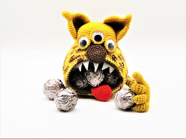 Gehäkeltes gelb-braunes Monster mit drei Augen, offenem Maul, roter Zunge und silbernen Pralinen.
