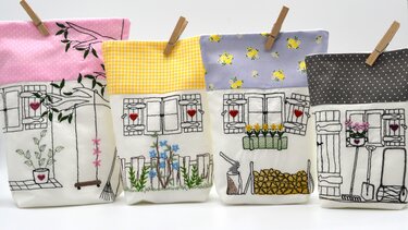 Vier bestickte Stoffhüllen im Landhausstil, jede mit Fenster- und Gartenmotiv, an Wäscheklammern befestigt.