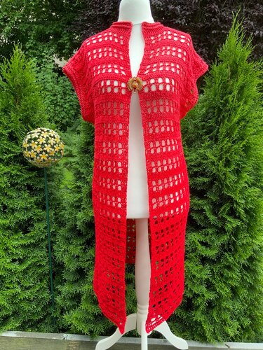 Knielange rote Häkelweste mit Reihen-Lochmuster, kurzem Ärmel und Brosche als Verschluss auf Schaufensterpuppe vor grünen Hecken