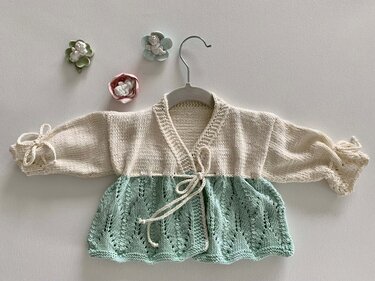 Zweifarbige gestrickte Baby-Wickeljacke in Creme und Mintgrün mit Lochmuster und Bindeband auf einem Kleiderbügel