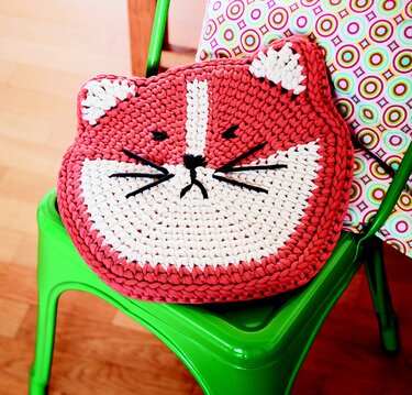 Gehäkeltes Katzenkopfkissen in Terrakotta und Weiß mit schwarzen Schnurrhaaren auf einem grünen Stuhl