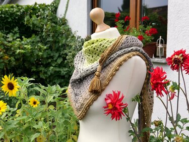 Gehäkeltes Dreieckstuch in Grün, Grau und Beige mit Lochmuster und Quaste auf einer Schneiderpuppe vor Gartenblumen.