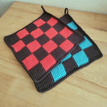 Zwei gehäkelte Topflappen mit Karomuster in Dunkelbraun, Rot und Türkis auf Holzoberfläche.