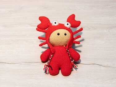 Gehäkelte Amigurumi-Krabbe in rotem Kostüm auf hellem Holzhintergrund.