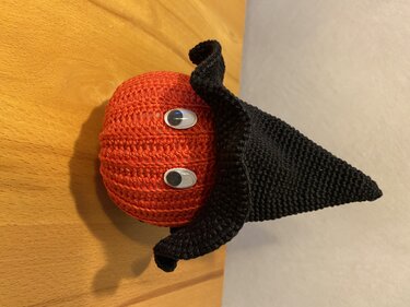 Gehäkelter orangefarbener Kürbis mit schwarzem spitzem Hexenhut und Wackelaugen