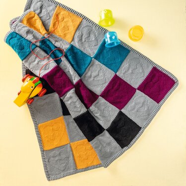 Gestrickte Babydecke mit karierten Quadraten und eingestrickten Herzmotiven, daneben Holzziehspielzeug und bunte Becher