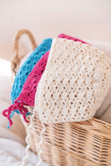 Drei gehäkelte Babymützen in Creme, Fuchsia und Türkis mit Lochmuster und Bindebändern, liegend in einem Weidenkorb.