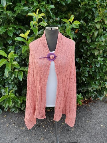 Korallfarbenes gestricktes Umschlagtuch mit Lochmuster, befestigt mit violetter Brosche vor grünem Laub