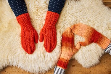 Zwei Paare gestrickter Handschuhe mit Zopfmuster: orangefarbene Fäustlinge und farbverlaufende Fingerhandschuhe auf hellem, flauschigem Untergrund.