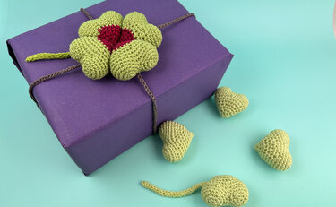 Violettes Geschenk mit einem grünen gehäkelten Kleeblatt und mehreren kleinen gehäkelten Herzen auf türkisfarbenem Untergrund