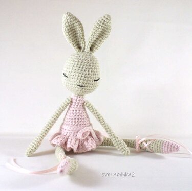 Light green crocheted amigurumi bunny wearing a pink ruffled ballet dress with ribbon-tied ankles, seated on a white background.