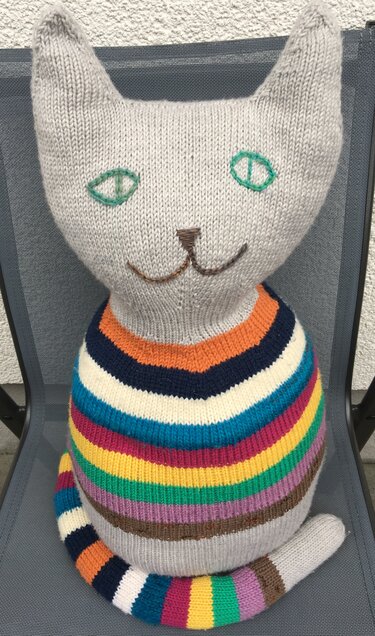 Gestrickte Amigurumi-Katze mit hellgrauem Kopf und bunten, horizontalen Streifen am Körper und Schwanz, sitzt auf einem Stuhl.
