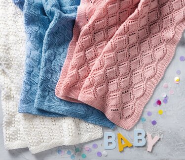 Drei gestrickte Babydecken in Weiß, Blau und Rosa mit Ajour-, Diamant- und Strukturmustern, daneben Holzbuchstaben 'BABY' und Konfetti