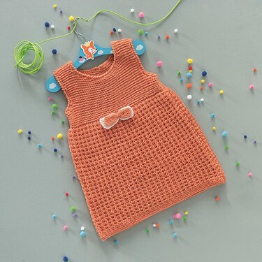 Korallfarbenes gestricktes Babykleid mit Schleife auf blauem Kinderbügel, umgeben von bunten Pompons