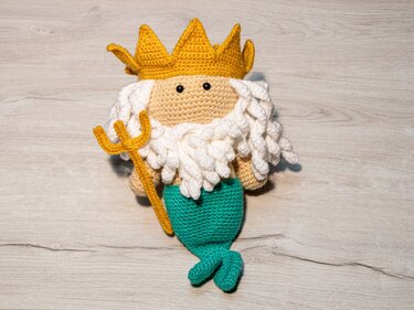 Gehäkelter Amigurumi-Meereskönig mit goldgelber Krone, weißem Bart, türkisgrüner Flosse und Dreizack.