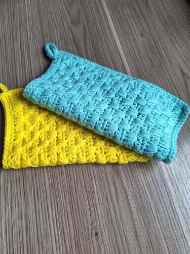 Zwei gehäkelte Topflappen, gelb und türkis, mit Strukturmuster und Aufhängeschlaufen auf Holzuntergrund.