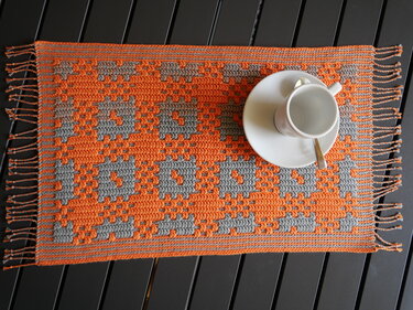 Oranger und grauer gehäkelter Tischläufer mit geometrischem Muster, Fransen und einer weißen Tasse auf Untertasse