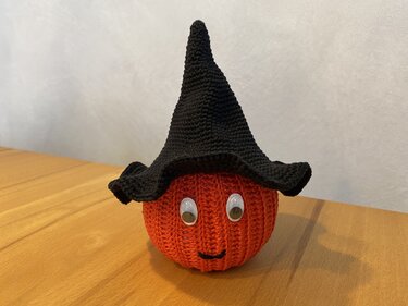 Gehäkelter orangefarbener Kürbis mit Kulleraugen, gesticktem Lächeln und schwarzem Hexenhut auf einem Holztisch.