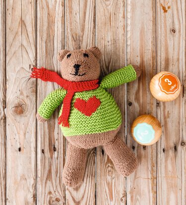 Gestrickter brauner Teddybär mit grünem Pullover, rotem Herz und rotem Schal auf Holz