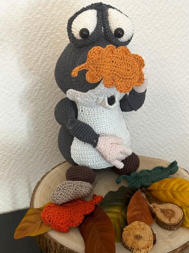 Gehäkelter Amigurumi-Maulwurf in Grau und Weiß hält ein orangefarbenes Blatt vor dem Gesicht; auf einer Holzscheibe mit herbstlichen Blättern arrangiert.