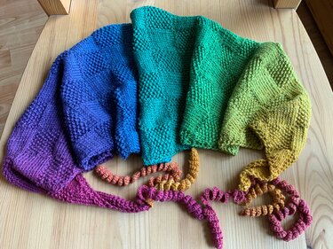 Gestricktes Dreieckstuch mit Regenbogen-Farbverlauf und spiralförmigen Borten auf Holz