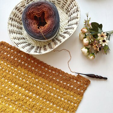Overhead view of a partially crocheted rectangular shawl in yellow-to-brown gradient yarn, a yarn cake in a woven basket, and a wooden crochet hook.