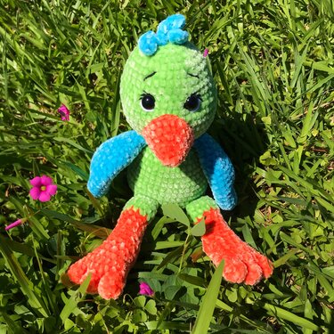 Gehäkelter Amigurumi-Vogel in Grün, Blau und Orange, sitzend im Gras mit kleinen lila Blüten