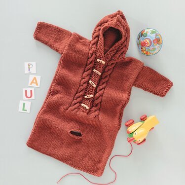 Rostroter gestrickter Babyschlafsack mit Kapuze, Zopfmuster und Holzknöpfen, liegend neben Holzspielzeug