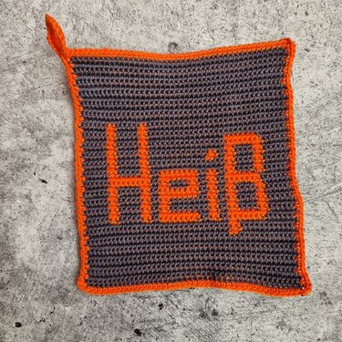 Gehäkelter grauer Topflappen mit orangefarbener Umrandung und dem Schriftzug „Heiß“, mit Aufhängeschlaufe