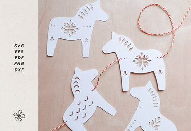 Vier weiße, ausgestanzte Pferde-Anhänger aus Papier mit dekorativen Musterausschnitten und rot-weißem Garn auf hellem Holzuntergrund