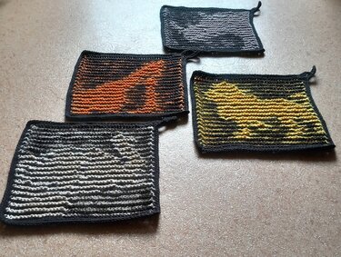Vier gestrickte Topflappen mit kontrastreichen Tierumrissen in Orange, Gelb, Hellgrau und Beige auf dunklem Grund, alle mit dunklem Rand und Aufhängeschlaufen.