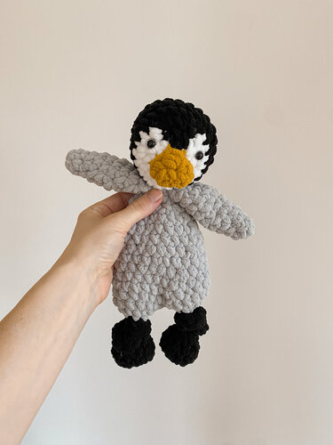 Hand hält kleines gehäkeltes Pinguin-Schmusetuch mit grauem Körper, schwarzem Kopf und gelbem Schnabel