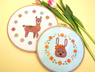 Zwei Kreuzstichbilder in Holzrahmen: Lama auf weißem Stoff und Hasenkopf auf hellblauem Stoff, daneben rosa Tulpen auf gelbem Hintergrund.