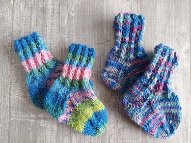 Zwei Paare bunter, handgestrickter Babysocken auf hellem Holzuntergrund, eines gestreift, das andere meliert.