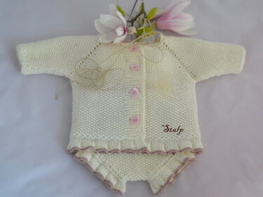 Cremefarbene gestrickte Babyjacke mit passendem Windelhöschen, rosa Knöpfen und Wellenkante, liegend auf hellem Hintergrund