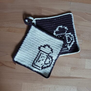 Zwei quadratische gehäkelte Topflappen in Creme und Dunkelbraun mit Bierkrug-Motiv und Aufhängeschlaufe auf Holzuntergrund