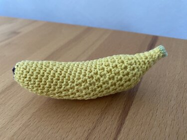 Gelbe gehäkelte Banane mit grünem Stiel und dunkler Spitze auf hellem Holztisch.