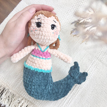 Hand hält gehäkelte Amigurumi-Meerjungfrau mit braunem Haar, rosa Oberteil und türkisfarbenem Schwanz auf hellem Hintergrund