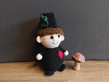 Gehäkelte Amigurumi-Puppe: Kobold mit schwarzem Zylinder, grünem Kleeblatt, braunen Haaren und rotem Herz, neben einem Holzpilz.