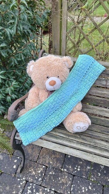 Gehäkelter blau-grüner Loop-Schal auf Holzbank, drapiert über einem Teddybär