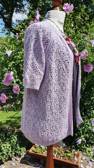Helllilafarbene offene Strickjacke mit Ajourmuster auf Schneiderpuppe vor blühenden Sträuchern