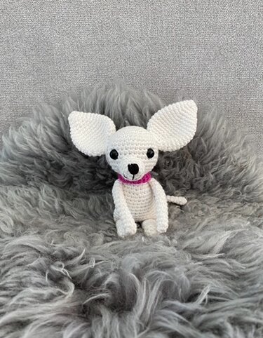 Sitzender weißer Amigurumi-Chihuahua mit rosa Halsband auf grauem Kunstfell