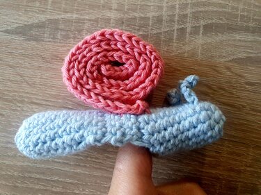 Gehäkelte kleine Schnecke in Hellblau mit rosa Schneckenhaus auf einem Finger