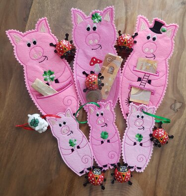 Mehrere rosa Filz‑Glücksschweine als ITH‑Stickmotive, bestickt mit Kleeblättern, Pilzen und Zylindern; teils mit Geldscheinen und Münzen in Taschen, arrangiert auf Holzuntergrund.