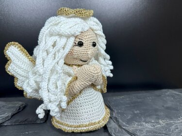 Gehäkelte Engelsfigur in Weiß mit goldenen Rändern, Heiligenschein und gefalteten Händen vor dunklem Hintergrund