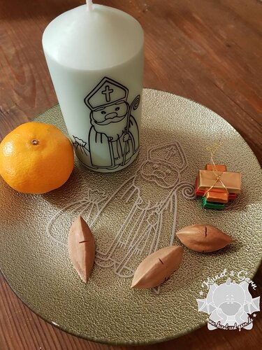 Weiße Stumpenkerze mit schwarzem Nikolaus-Plott auf goldfarbenem Teller mit Mandarine, Nüssen und kleinem Geschenk.