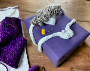 Violett verpacktes Geschenk mit gerüschter Häkel-Schleife, weißen Bändern und kleiner gelber Blüte