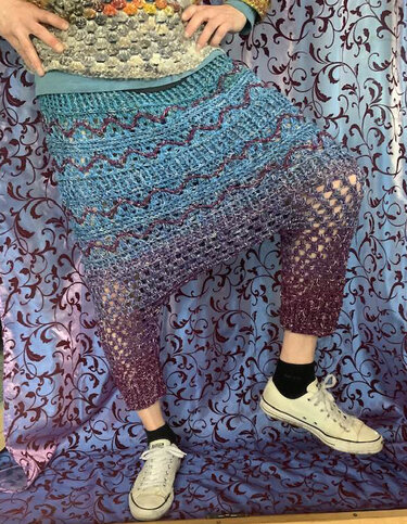 Nahaufnahme einer gehäkelten Hose mit blau-lila Farbverlauf, Wellenreihen und offenem Netzmuster an den Unterschenkeln.