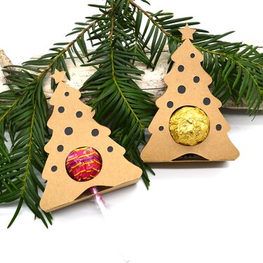 Zwei Tannenbaum-Falzboxen aus Kraftkarton mit runder Öffnung, jeweils gefüllt mit einem Lolli und einer Schokoladenkugel, auf weißem Untergrund neben Tannenzweigen.